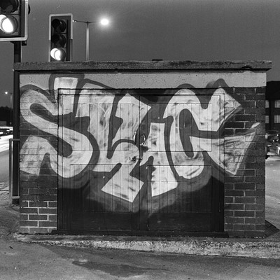 The image is a black and white photo of a brick building with graffiti on it. There are several traffic lights visible in the scene, one near the top left corner, another at the top right corner, and two more towards the bottom right side. A car can be seen parked next to the building, and there's also a person standing close to the center of the image.