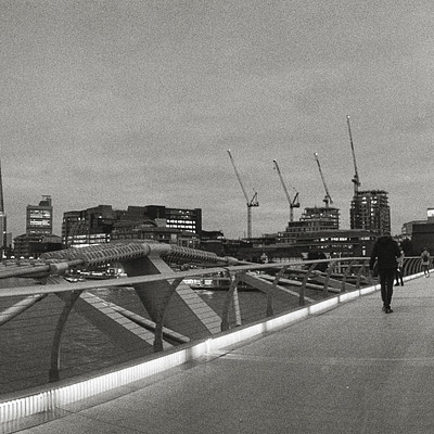 The image is a black and white photo of a bridge overlooking a city. There are several people walking on the bridge, with some closer to the foreground and others further away. A few individuals can be seen carrying handbags as they cross the bridge.