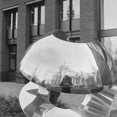 The image is a black and white photo of a large, reflective metal sculpture in the foreground. Behind it, there are buildings visible, including one with a clock tower. A person can be seen standing near the sculpture, possibly admiring its unique design.