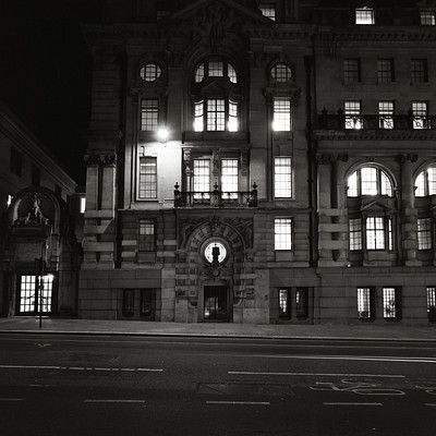 The image is a black and white photo of an old building with many windows. It appears to be a large, historic structure that has been well-maintained over the years. There are several people in the scene, some standing near the entrance while others are scattered throughout the area.