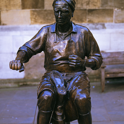The image features a statue of a man sitting on a bench, possibly made from metal. He is wearing a hat and appears to be holding a baseball bat in his hand. The statue is positioned outdoors, with the bench visible behind it.