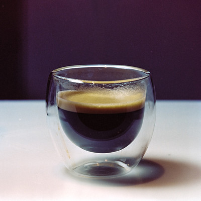 The image features a close-up of a glass cup filled with coffee, placed on a table. The cup is positioned in the center of the scene and appears to be empty, except for some foam at the top. The focus of the photo is on the glass cup, highlighting its contents and texture.