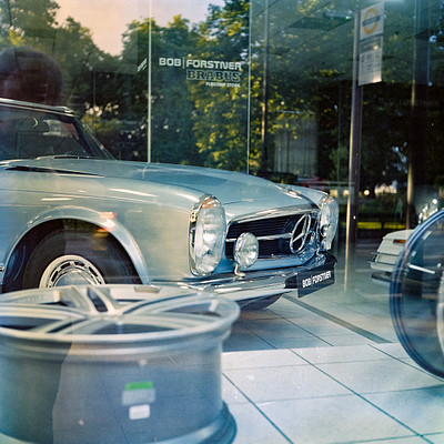 The image features a silver car parked in front of a building, possibly a dealership or showroom. The car is positioned near the center of the scene and appears to be an older model. There are several tires visible around the car, with some placed closer to the left side and others on the right side.