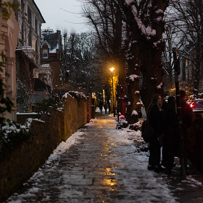 The image depicts a snowy street lined with trees, where people are walking down the sidewalk. There is a couple of individuals standing on the sidewalk, and others can be seen further down the path. A car is parked at the end of the street, adding to the winter atmosphere.