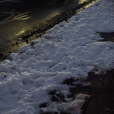 The image features a sidewalk with a pile of snow on it. The snow is scattered all over the ground, creating a wintery atmosphere. There are also some ice patches visible in the scene.
