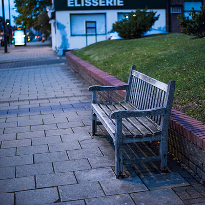 The image features a lonely park bench situated on the sidewalk near a brick wall. It is located in front of a building, possibly an old bakery or cafe, as indicated by the presence of a sign above it. There are several cars parked nearby, with one car closer to the left edge of the image and two others further back on the right side.