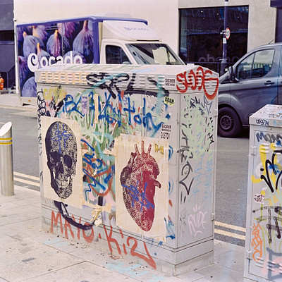 The image features a graffiti-covered box on the side of a street. The box is covered in various colors and designs, including skulls and hearts. A truck can be seen parked nearby, adding to the urban atmosphere of the scene.