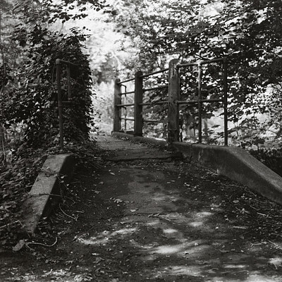 The image is a black and white photo of an old stone bridge with a metal gate. The bridge has a dirt road leading to it, and the gate appears to be closed. There are trees in the background, adding to the overall atmosphere of the scene.