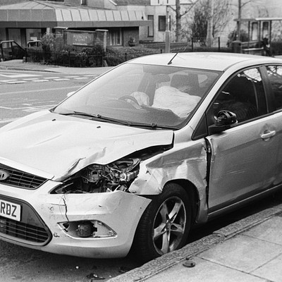 The image is a black and white photo of a car that has been in an accident. It appears to be a Ford, as indicated by the license plate. The front end of the vehicle is severely damaged with a broken windshield and a crushed hood.