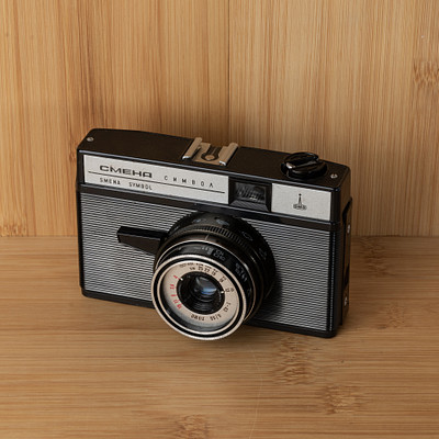 The image features an old-fashioned camera sitting on a wooden table. The camera is black and white, with a large lens in the front. It appears to be an antique model, possibly from the 1950s or earlier.