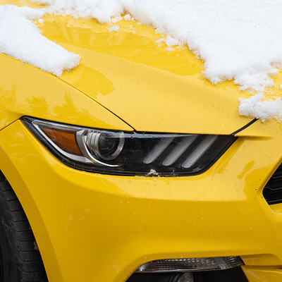 The image features a yellow Mustang car with its hood up, covered in snow. The car is parked on the street and appears to be well-maintained. The snow covers not only the hood but also the windshield, giving the car a unique winter appearance.