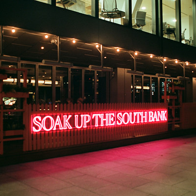 The image features a restaurant with a large sign that reads "soak up the south bank" in red neon lights. The sign is located outside of the building, and there are several chairs placed around the area. Inside the restaurant, numerous dining tables can be seen, along with various potted plants scattered throughout the space.
