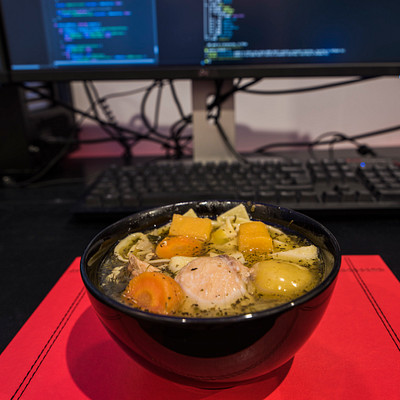The image features a desk with two computer monitors on it. A bowl of soup is placed in the center of the desk, surrounded by various items such as a keyboard and a mouse. There are also several carrots scattered around the bowl, adding to the visual appeal of the scene.