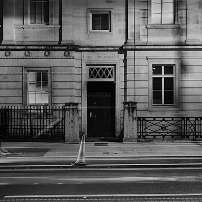 The image is a black and white photo of an old building with a large doorway. The building has a stone facade, giving it an antique appearance. There are several windows on the building, some of which have bars covering them.
