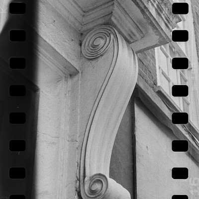 The image is a black and white photo of an ornate decorative piece, possibly a carved stone or marble column, located on the side of a building. It appears to be part of a window frame, with a window visible in the background. The column has a scroll design at its base, adding to its intricate appearance.