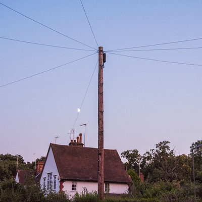 The image features a white house with a red roof, situated in the middle of a field. A large wooden pole is standing next to the house, and there are power lines running above it. In the background, there is a beautiful view of the moon rising over the horizon.