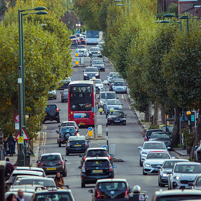 The image depicts a busy city street filled with traffic, including cars and buses. There are several cars of various sizes scattered throughout the scene, some driving down the road while others are parked on the side. A red double-decker bus is also present in the middle of the street, adding to the bustling atmosphere.