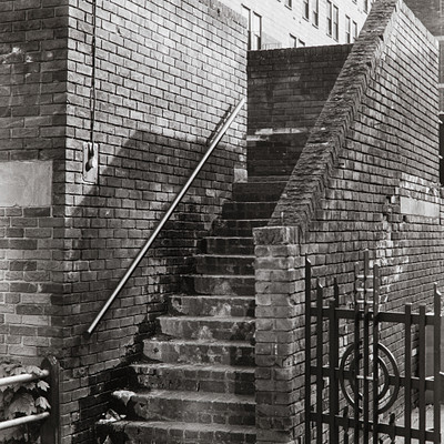The image is a black and white photo of an old brick building with two sets of stairs leading up to the second floor. The staircase appears to be made of bricks, giving it a classic appearance. There are several windows on the building, some located near the top of the stairs while others can be seen further down the side of the structure.