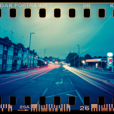 The image is a black and white photo of a busy street at night. There are several cars driving down the road, with some appearing blurry due to motion. A few traffic lights can be seen along the street, helping control the flow of vehicles.
