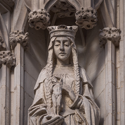 The image features a statue of a woman, possibly Mary or another religious figure, holding a baby in her arms. She is wearing a crown and appears to be the central focus of the scene. The statue is located near an archway with a stone wall behind it.