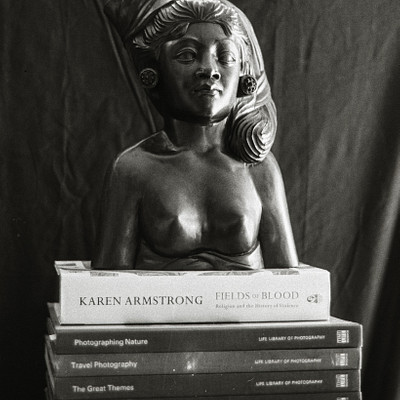 The image features a statue of a woman sitting on top of several books. The statue is positioned in the center of the scene, with the books surrounding it. There are at least nine books visible in the picture, some stacked vertically and others horizontally.