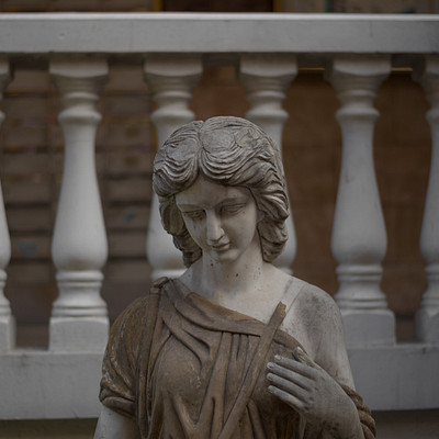 The image features a statue of a woman, possibly an angel or a goddess, with her hands on her chest. She is standing in front of a white railing and appears to be looking downward. The statue has a classical appearance, giving it a sense of elegance and grace.