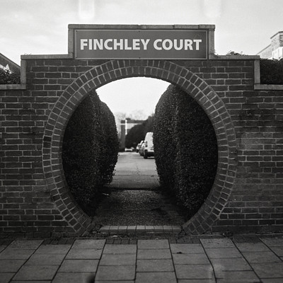 The image is a black and white photo of a brick archway with the words "Finchley Court" written above it. The archway leads to a street, where several cars are parked along the side. There are at least five cars visible in the scene, with some closer to the foreground and others further back.