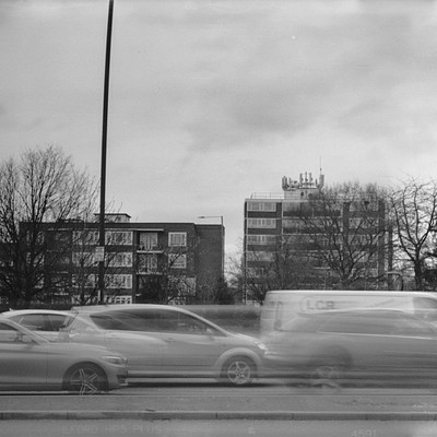 The image is a black and white photo of a busy city street filled with traffic. There are several cars driving down the road, including some that appear to be blurry due to motion. In addition to the cars, there are also two trucks visible on the street.