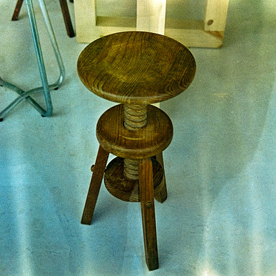 The image features a wooden table with three legs, placed on a white floor. The table is positioned in the center of the scene and appears to be made from wood. There are also two chairs visible in the background, one located near the left side of the room and another towards the right side.