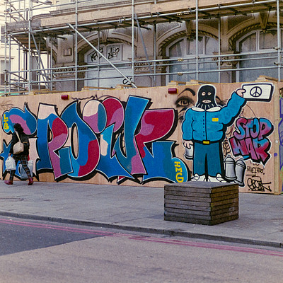 The image features a graffiti-covered wall with various writings and drawings on it. A large mural of a man is also present, adding to the artistic display. In addition to the graffiti, there are several people scattered throughout the scene, some closer to the wall while others are further away.