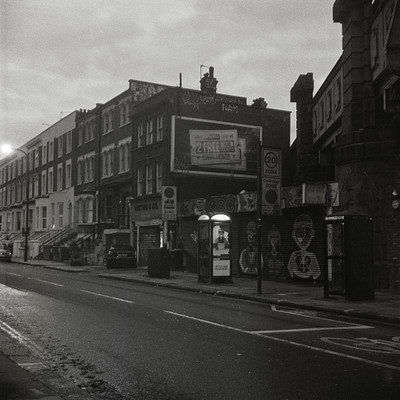 The image is a black and white photo of an urban street scene. There are several buildings on the side of the road, with one building featuring a large billboard. A bus stop can be seen in the middle of the scene, surrounded by various signs.