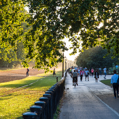 The image depicts a lively scene of people walking down a path near a park. There are numerous individuals, some carrying backpacks and handbags, enjoying their time outdoors. A group of people can be seen walking on the sidewalk next to a grassy area, while others are scattered throughout the scene.