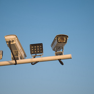 The image features a pole with several security cameras attached to it. There are five cameras in total, each mounted at different heights and angles on the pole. They appear to be surveying an area or monitoring traffic from above.