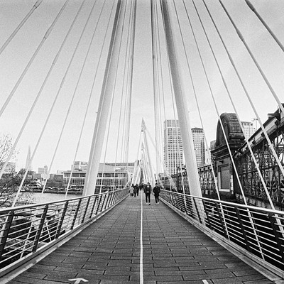 The image is a black and white photo of a bridge with people walking on it. There are three individuals visible in the scene, one near the center of the bridge, another closer to the left side, and the third person towards the right side. They appear to be enjoying their walk across the bridge.