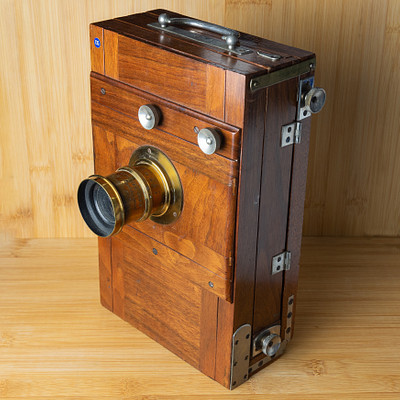 The image features a wooden box with a metal latch, which appears to be an old-fashioned camera. It has a large lens on the front and is placed on a table or countertop. The wooden box seems to have been repurposed as a display piece for this antique item.