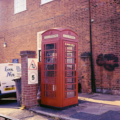 The image features a red telephone booth, which is an iconic symbol of London. It is located on the sidewalk next to a brick building and near a street corner. A truck can be seen parked in front of the phone booth, partially covering it from view.