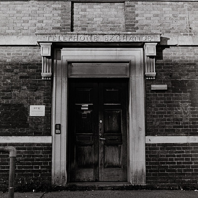 The image is a black and white photo of an old building with a large doorway. The doorway has a sign on it that reads "Telephone Exchange." The building appears to be made of brick, giving it a classic appearance.