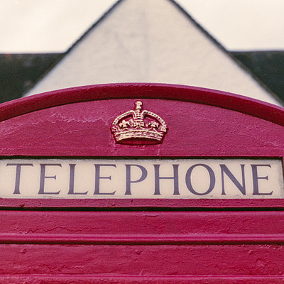 The image features a red telephone booth with the word "TELEPHONE" written on it. The booth is situated in front of a building, and there are two people visible in the scene - one person standing closer to the left side of the frame and another person further back on the right side.