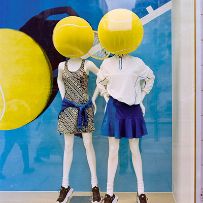 The image features a store display with two mannequins wearing tennis outfits. One of the mannequins is holding a tennis racket, while the other has a tennis ball on its head. Both are positioned in front of a blue and yellow background that adds to the overall tennis theme of the display.