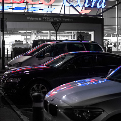The image is a black and white photo of several cars parked in front of a Tesco Extra store. There are five cars visible, with one car on the left side, two cars in the middle, and two more cars on the right side of the scene. In addition to the cars, there are three people present in the image.