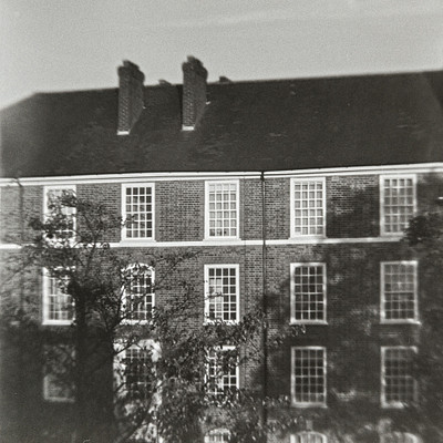 The image is a black and white photo of an old brick building with many windows. There are at least 14 visible windows, some of which have shutters on them. The building has a distinctive chimney on top, adding to its historical charm.
