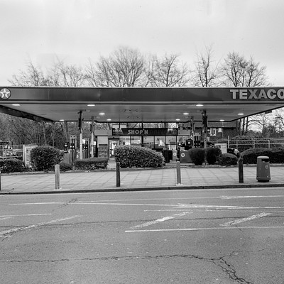 The image is a black and white photo of a gas station with the name "TEXACO" on top. There are several cars parked in front of the gas station, including one car to the left side, two cars in the middle, and another car further back on the right side. In addition to the vehicles, there is a person standing near the center of the scene, possibly attending to their vehicle or waiting for someone.