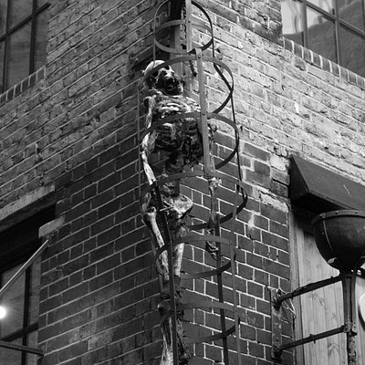 The image features a skeleton figure, possibly made of bones or metal, hanging from the side of a brick building. It appears to be an artistic display or decoration on the wall. The scene is in black and white, giving it a classic feel.