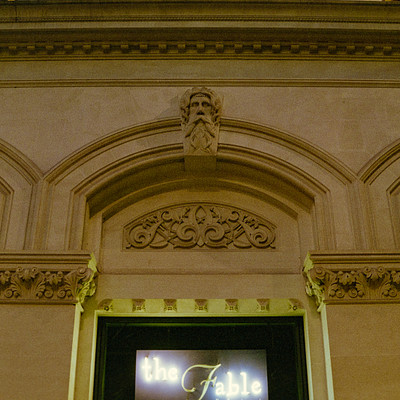 The image features a large building with ornate stonework and carved decorations. Above the entrance, there is a sign that reads "The Fable." The building has an old-fashioned appearance, giving it a sense of history and charm.