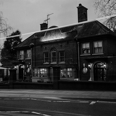 The image is a black and white photo of an old building, possibly a hotel or a restaurant. It has a brick facade with a gable roof, giving it a classic appearance. There are several windows on the building, some of which have lights shining through them.
