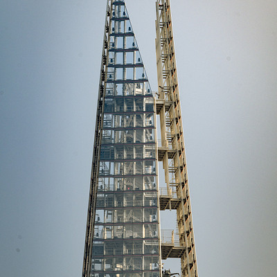 The image features a tall, thin glass building with many windows. It appears to be an observation tower or a skyscraper, as it is situated in the middle of a cityscape. The building has a unique design and stands out among other structures in the area.