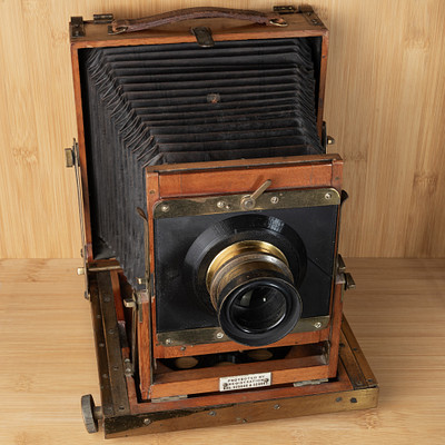 The image features an old, antique camera sitting on a wooden table. It is an old-fashioned camera with a large lens and a black frame. The camera appears to be in good condition despite its age, and it sits prominently on the table as if ready for use or display.