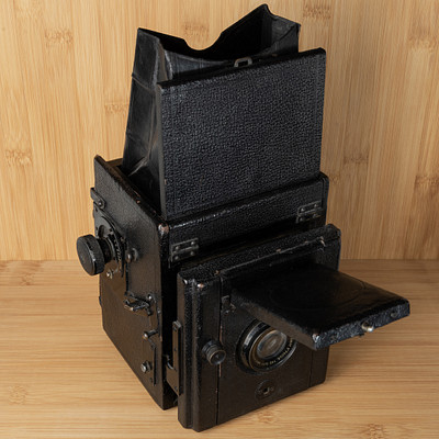 The image features an old, black camera sitting on a wooden table. It appears to be an antique or vintage model, possibly a box camera from the 1950s. The camera is open and ready for use, with its lens exposed.
