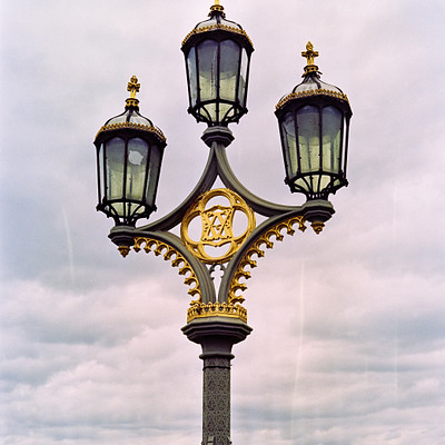The image features a large, ornate street light with four lights on top of it. The light is situated in the middle of a city and appears to be an old-fashioned design. In the background, there are several buildings visible, including one that has a clock tower.