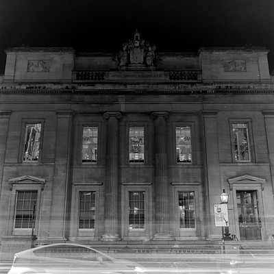 The image is a black and white photo of an old building, possibly a courthouse or a large house. It has many windows on the front side, with some of them being arched. A car is parked in front of the building, partially visible as it appears to be blurry due to motion.